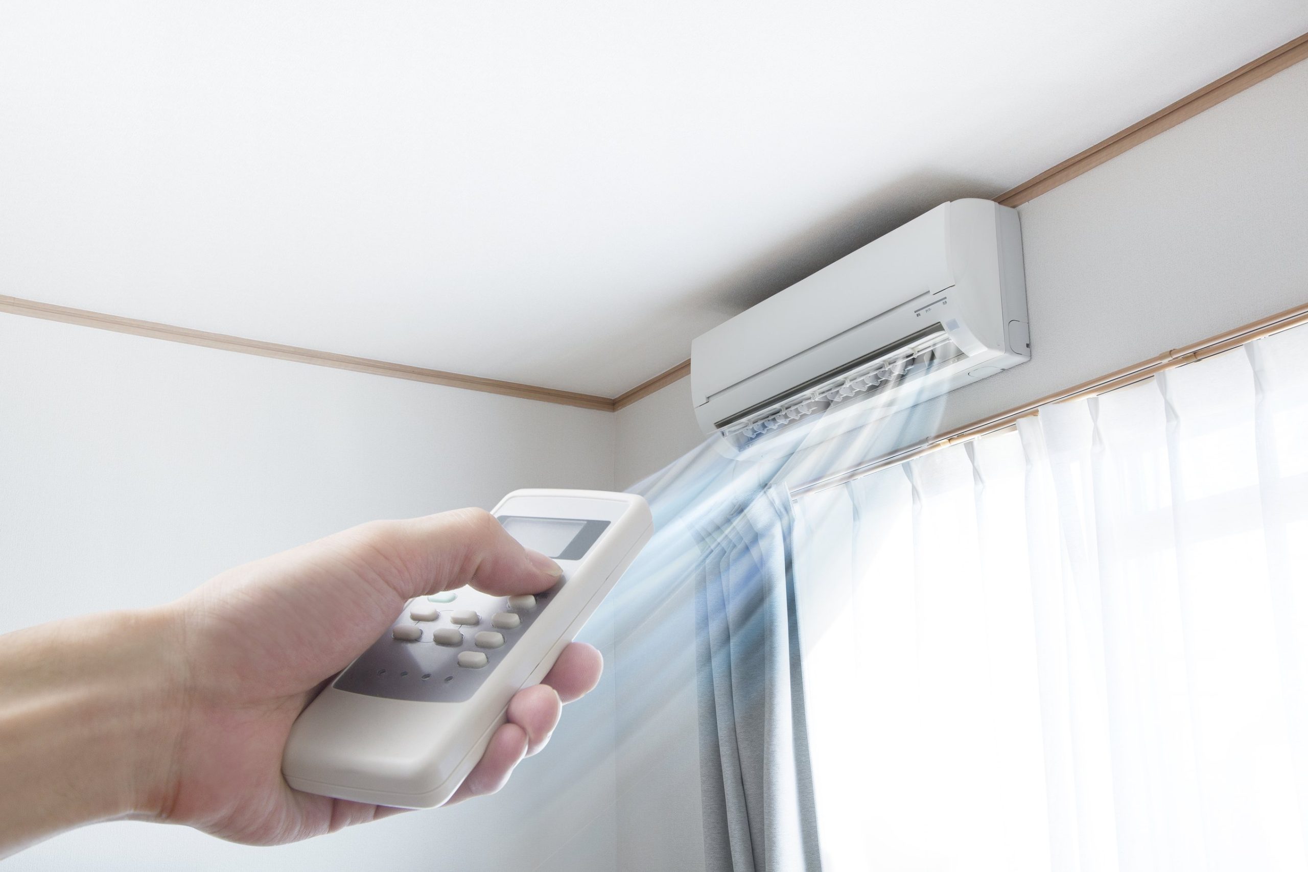 Hand holding remote control adjusting ductless mini-split air conditioner in a bright room with window curtains.